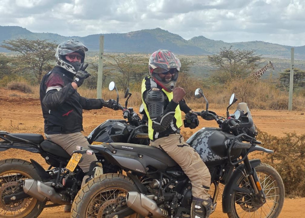 Two riders on adventure motorcycles with a giraffe behind them - Great Rift Kenya moto safari