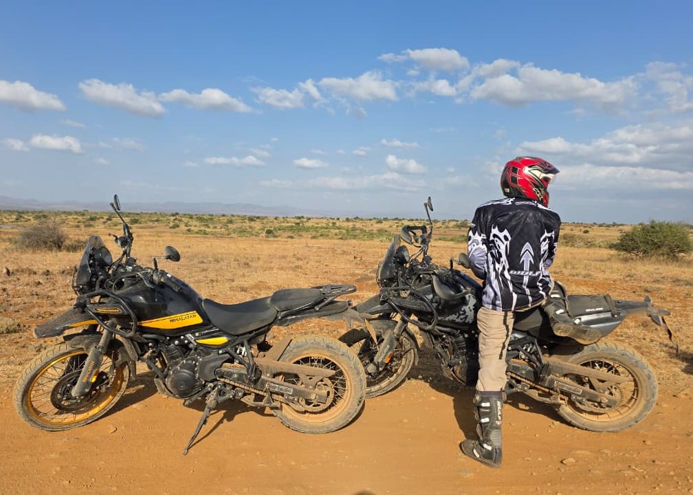 Rider and two motorcycles on red earth under a wide savannah sky